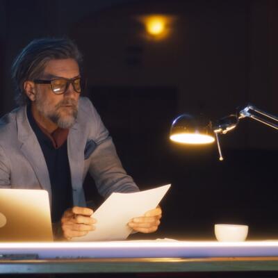 Lesender Mann mit Brille am Abend sitzt am Schreibtisch bei schlechter Beleuchtung.