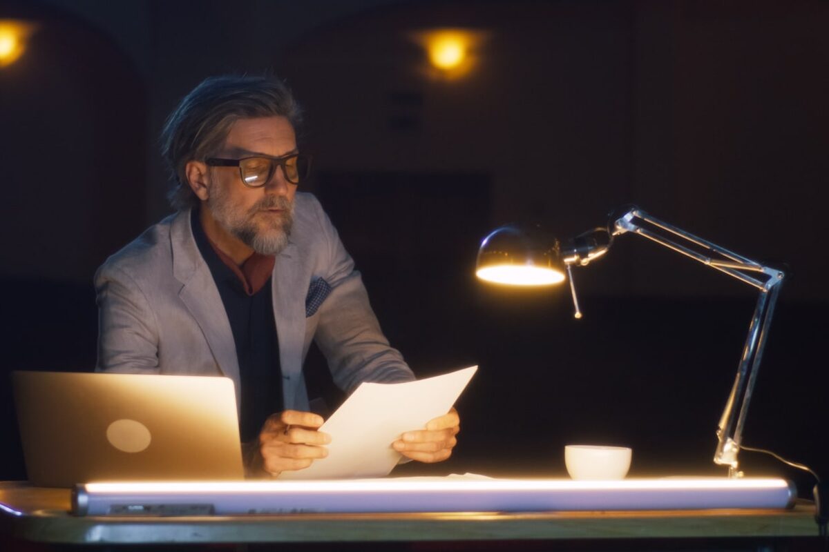 Mann mittleren Alters sitzt im Dunkeln am Schreibtisch mit heller Schreibtischlampe.