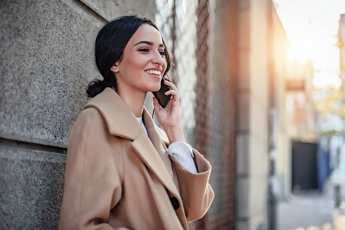 Junge Frau im beigen Mantel steht vor einer Häuserwand und telefoniert mit dem Handy.