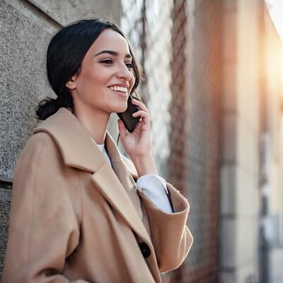 Junge Frau im beigen Mantel steht vor einer Häuserwand und telefoniert mit dem Handy.