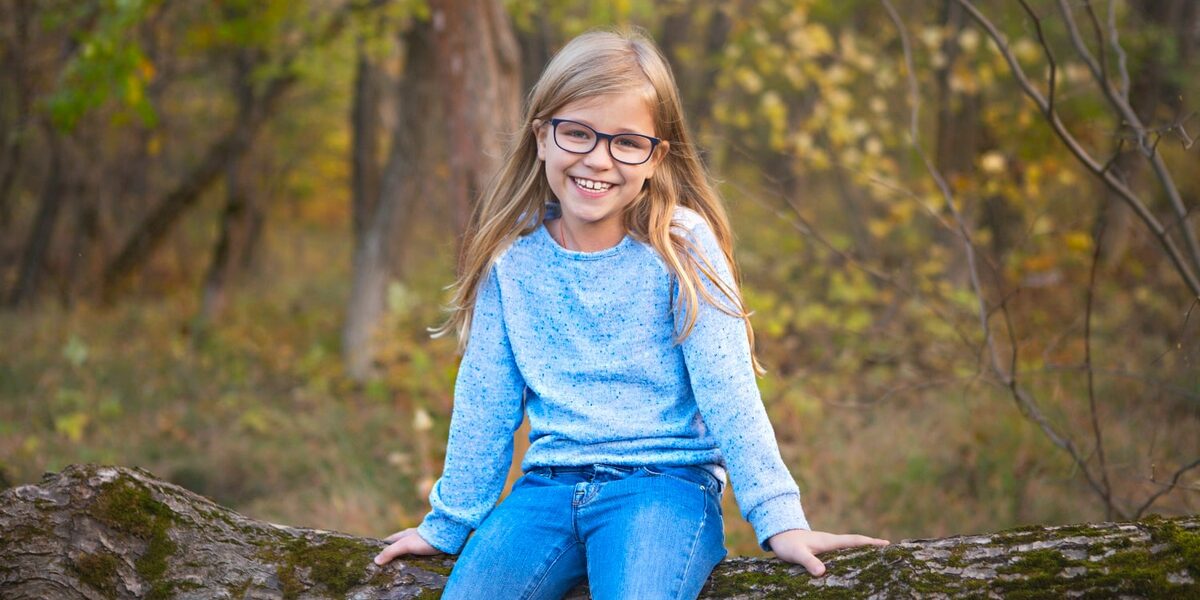 Mädchen mit Brille sitzt auf einem Baumstamm im Wald.